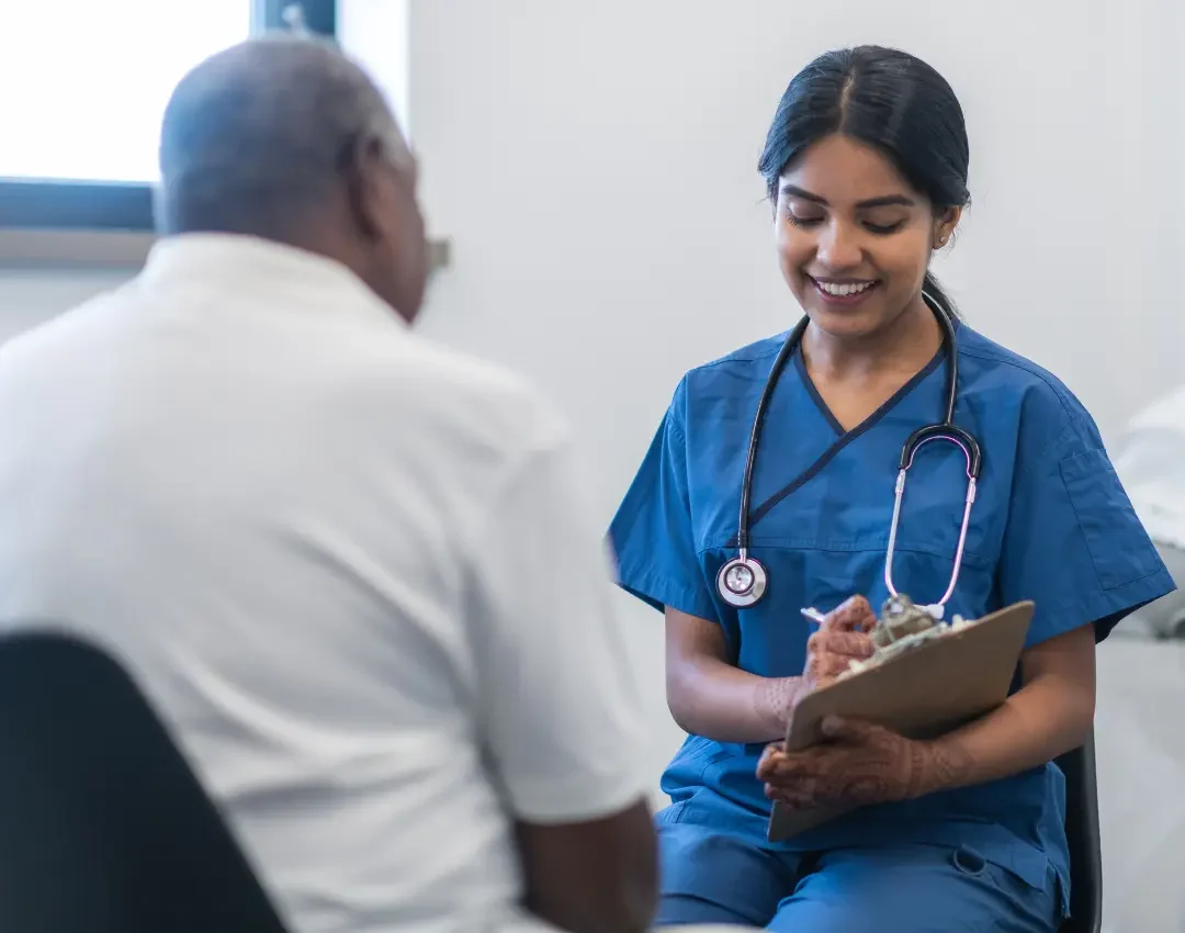 Medical professional in blue scrubs holding a clipboard while consulting with a patient.